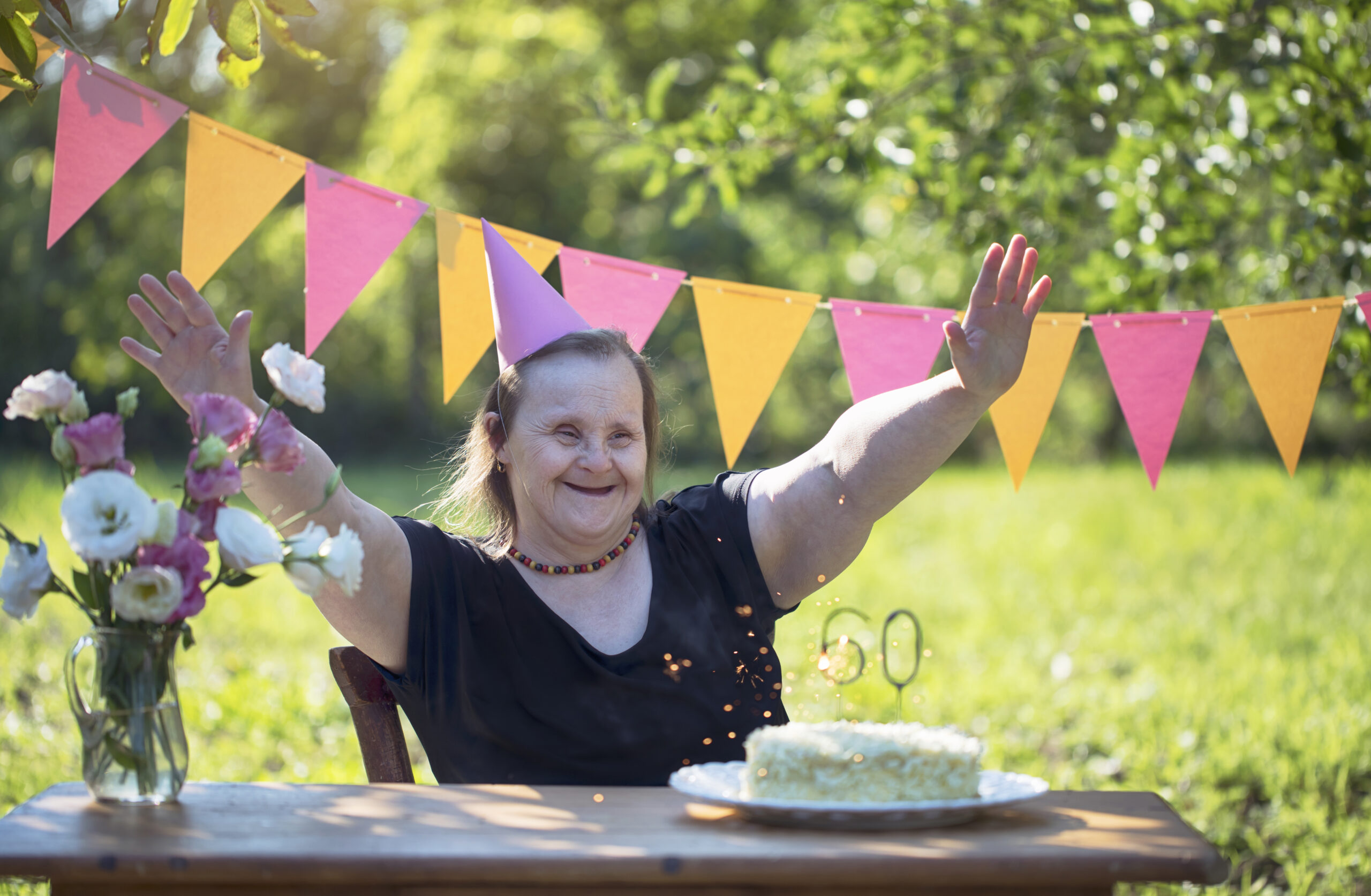 An adult living with an intellectual or developmental disability smiling with their hands in the air while sitting at a table with a birthday cake and birthday hat on.